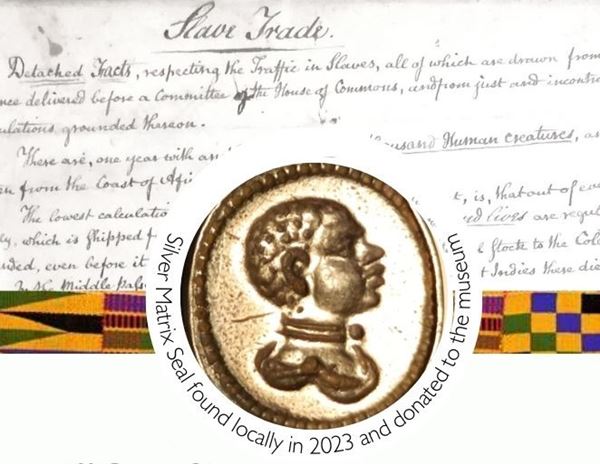 Photo of an oval silver seal depicting the head of a black person in profile, surrounded by the text 'Silver seal matrix found locally in 2023 and donated to the museums' In the background is a photo of a document discussing the slave trade, handwritten in black ink.